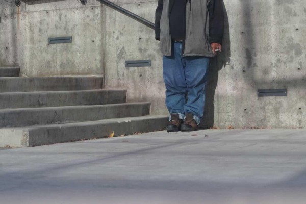 A man smokes a cigarette while leaning against a wall outside. Only his bottom half is shown and he is wearing boots, blue jeans and a grey jacket.