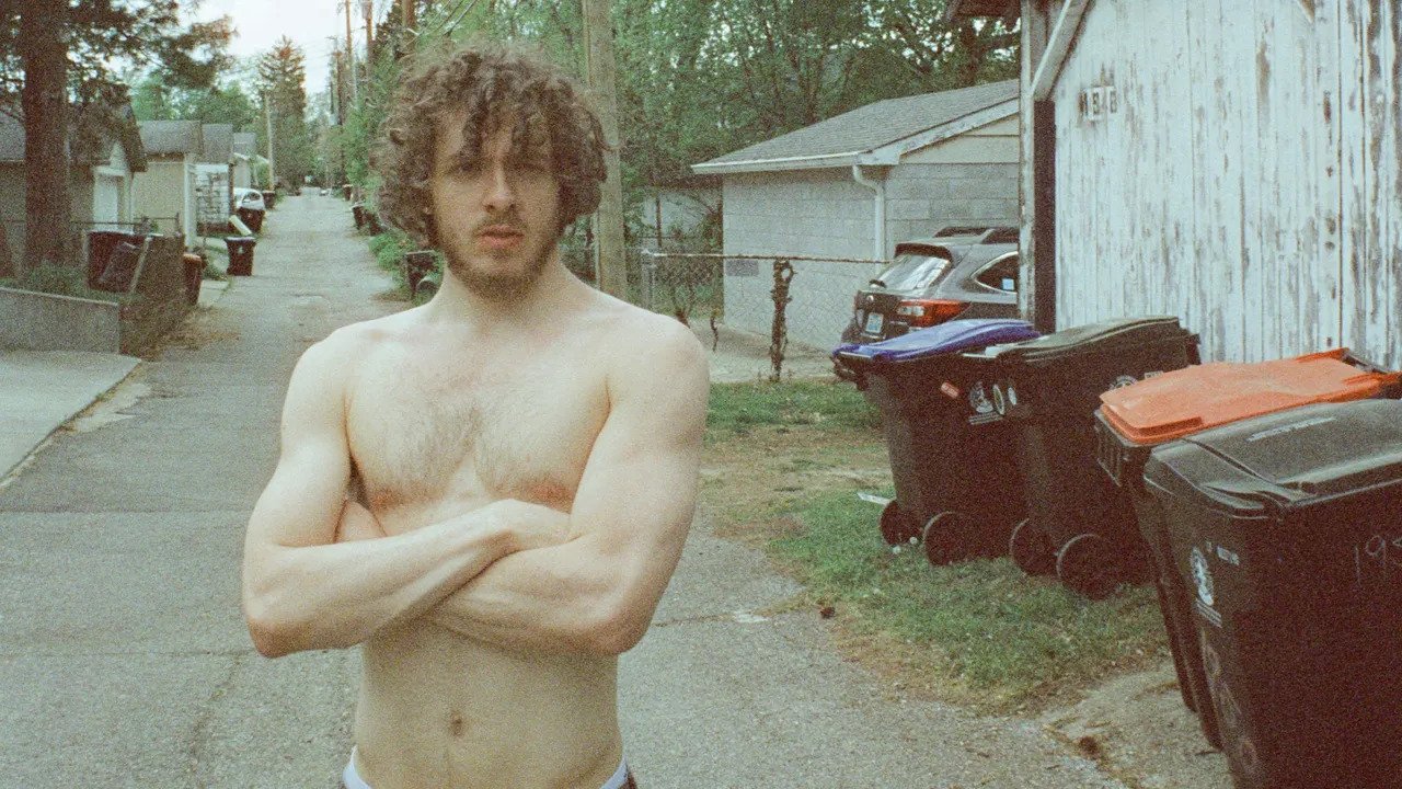 A photo of a man crossing his arms while standing shirtless in an alleyway